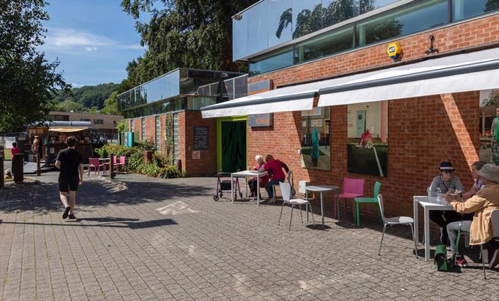 External angled view of gallery with awnings open and seating outside with visitors sitting underneath in the shade.